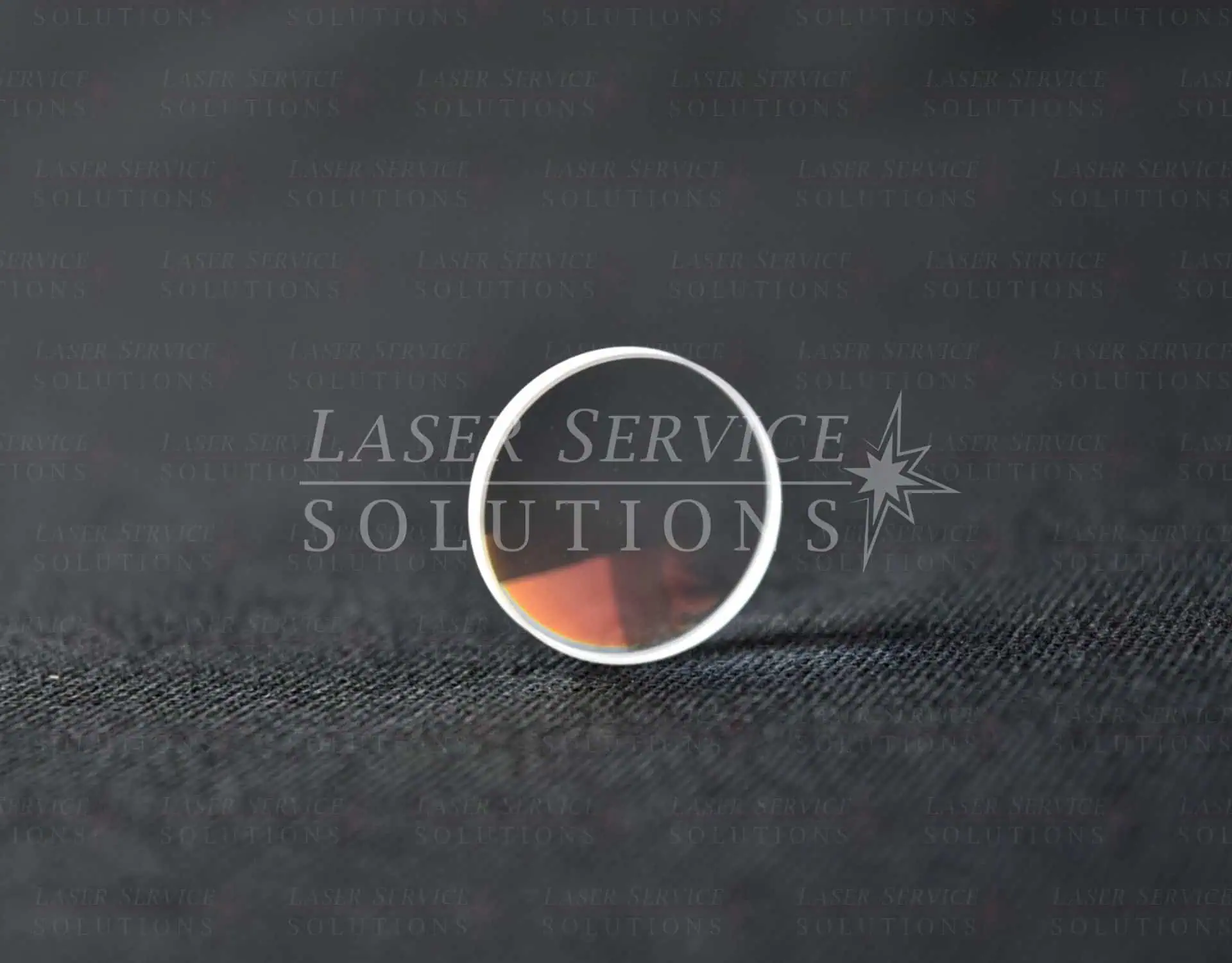 A round optical lens stands upright on a black textured surface, reflecting a subtle reddish-brown hue. The background is dark with faint Laser Service Solutions text watermark.