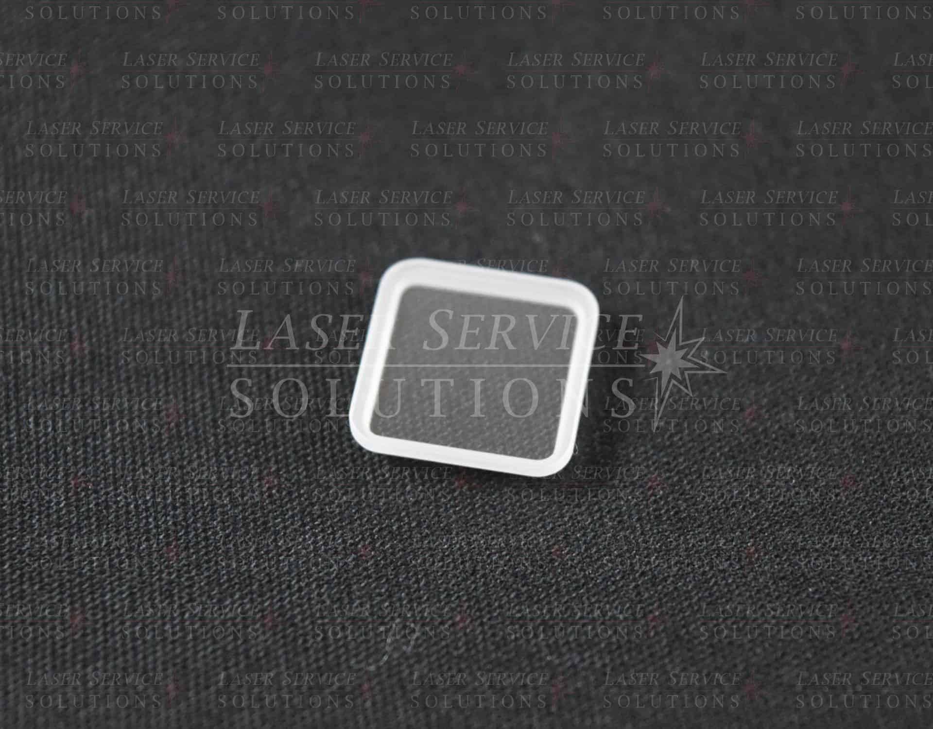 A Soprano XL Sapphire Handpiece Window, featuring a small, square, transparent glass design with white edges, is placed on a textured black surface.