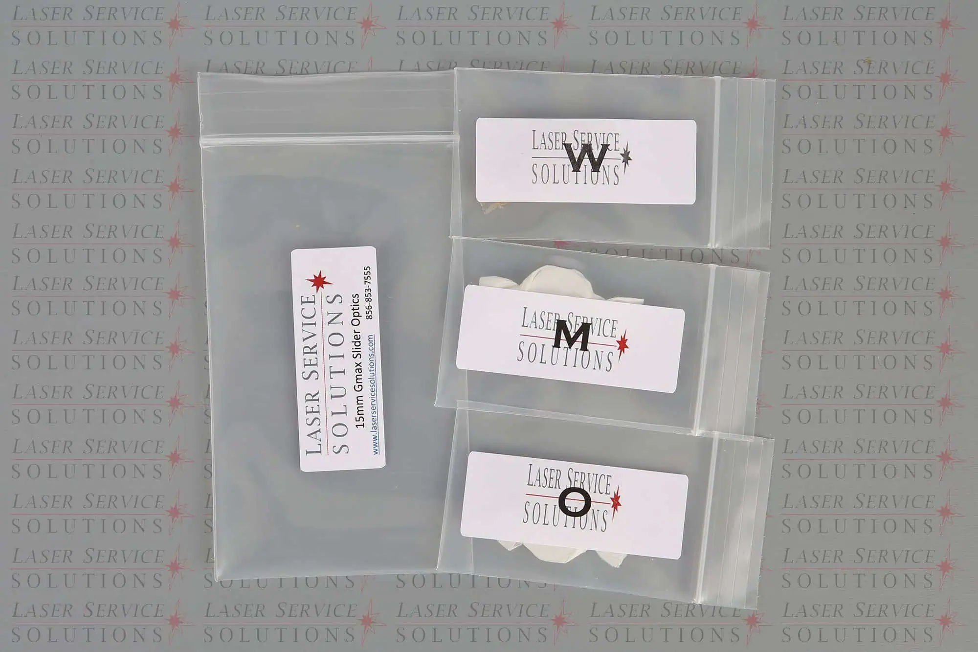 Four clear plastic bags, each labeled with Laser Service Solutions and a letter (W, M, N, O), contain components from the Candela Gmax Slider Optics Kits, all placed on a branded Laser Service Solutions background.