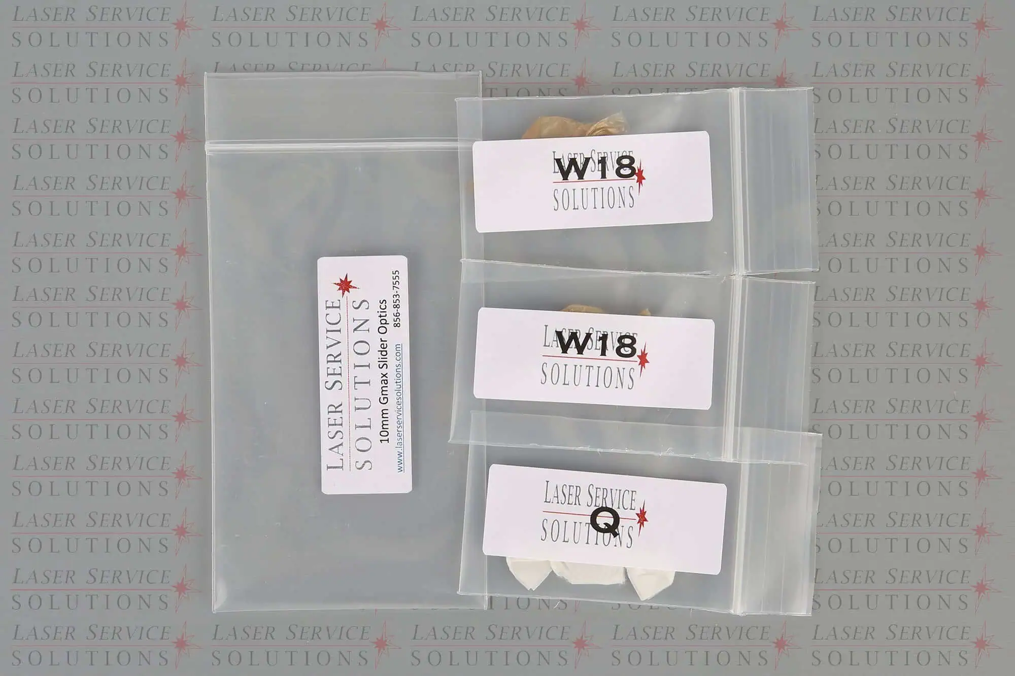 Four small transparent plastic bags labeled with Laser Service Solutions and various text, each containing components from the Candela Gmax Slider Optics Kits, are laid out on a background featuring a repeating Laser Service Solutions logo and pattern.