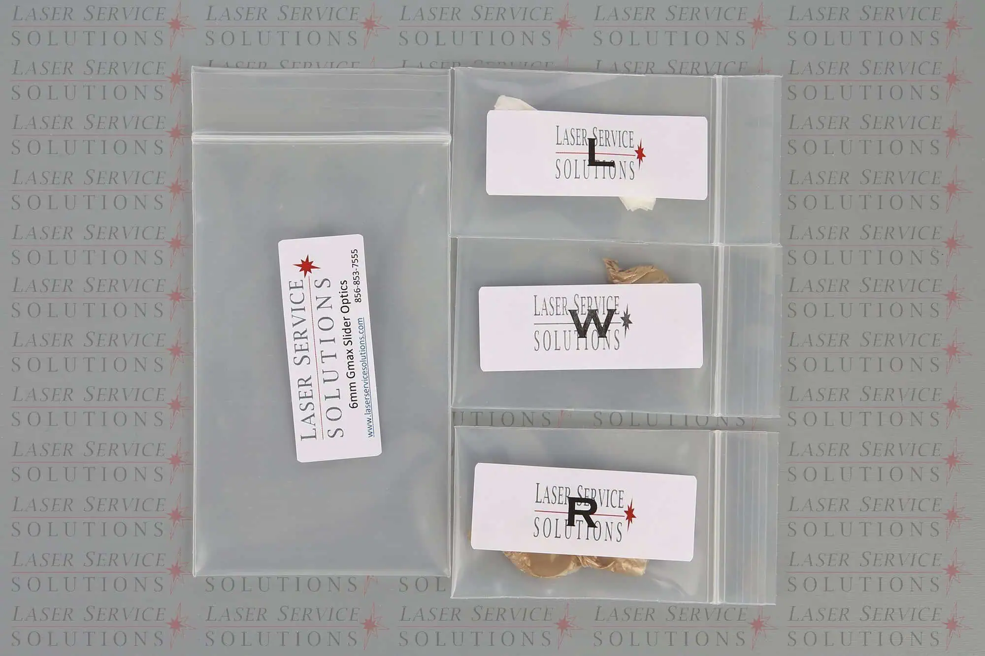 Four labeled plastic bags containing various small items from the Candela Gmax Slider Optics Kits, displayed on a surface featuring a repeating Laser Service Solutions logo and contact information in the background.