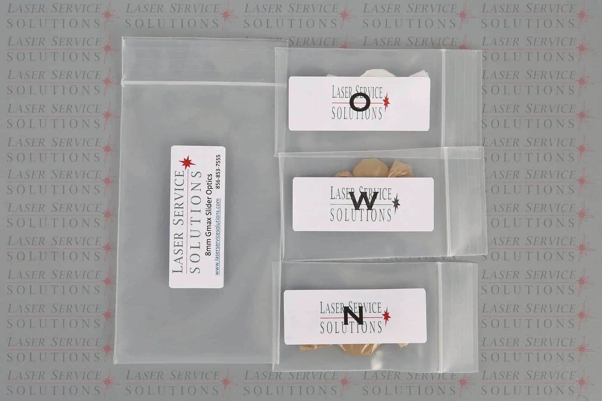 Four clear plastic bags are shown, three containing coins and labeled with “Laser Service Solutions.” The background has a repeating Laser Service Solutions logo. These are the Candela Gmax Slider Optics Kits.