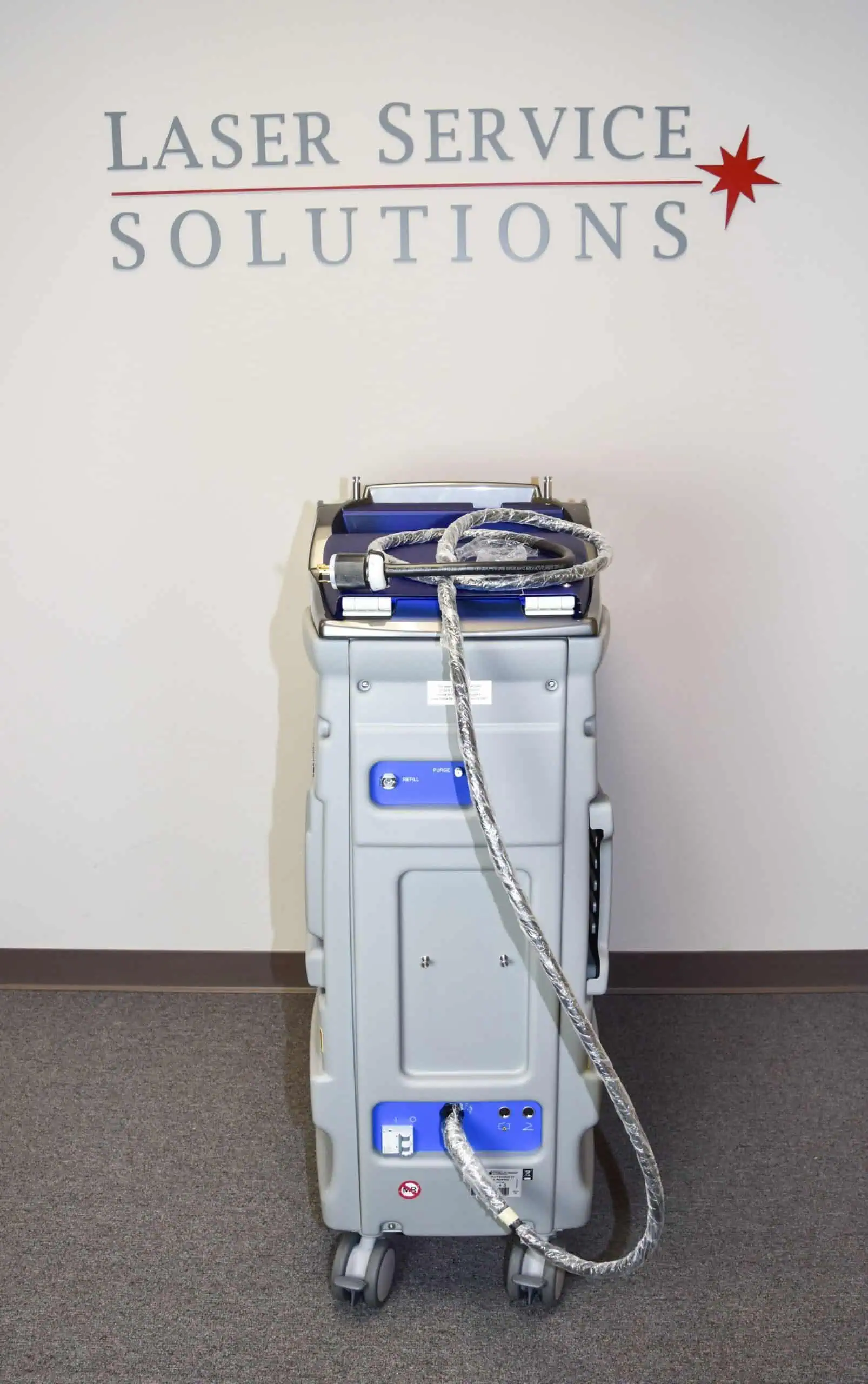 A DEKA Synchro Replay medical laser machine with attached cables stands on wheels in front of a wall labeled Laser Service Solutions with a red star symbol.