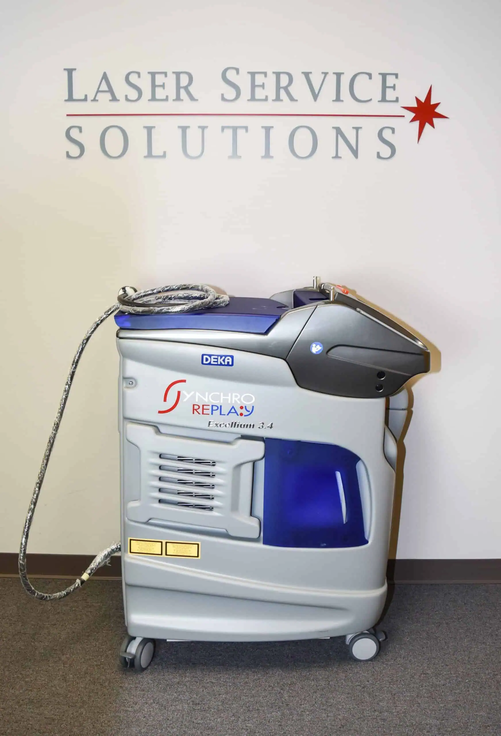 A DEKA Synchro Replay laser machine is positioned in front of a wall displaying the words Laser Service Solutions. The machine features a grey and blue design with a coiled cable on top and sits on a carpeted floor.
