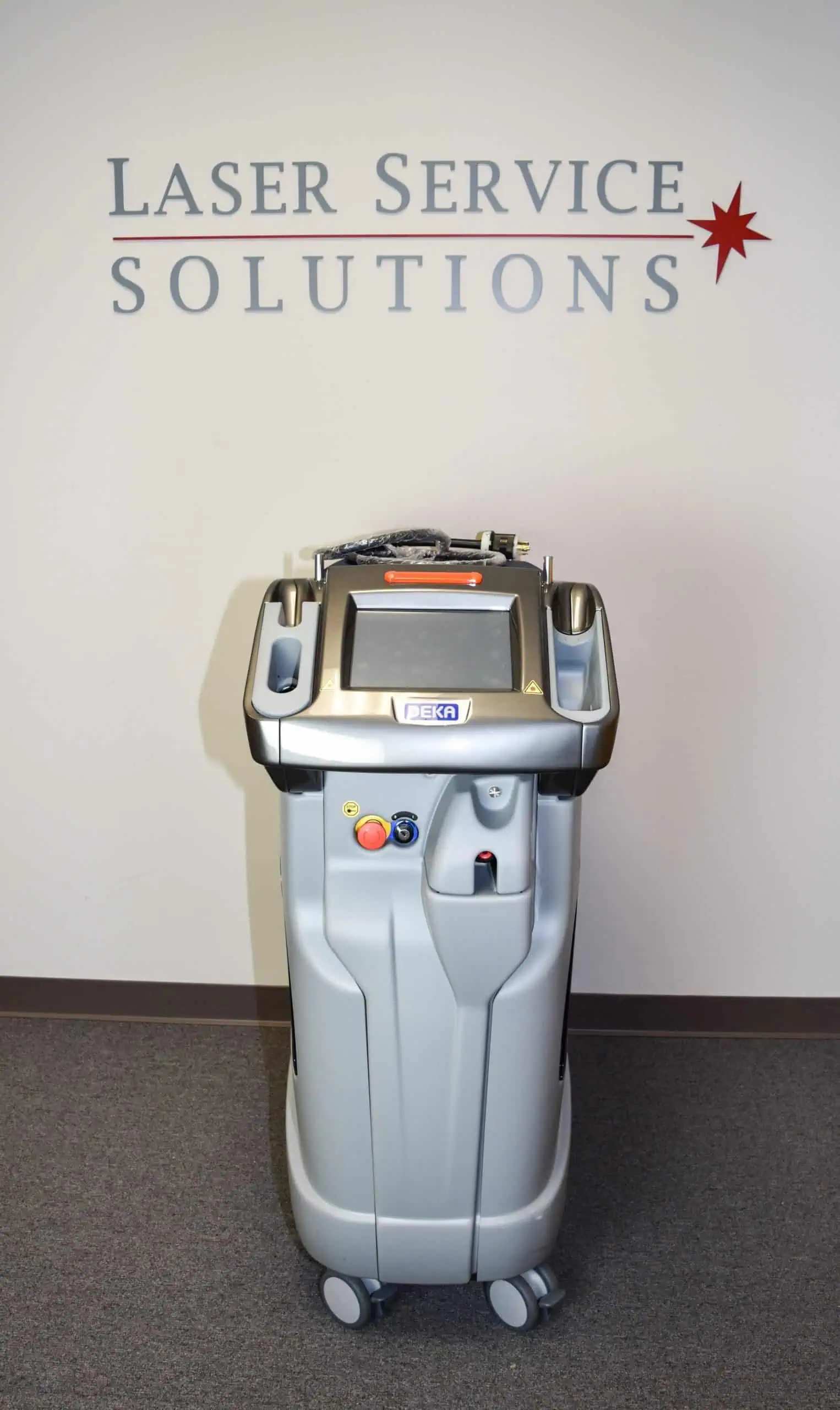 A DEKA Synchro Replay medical laser machine on wheels is positioned in front of a wall with the text Laser Service Solutions and a red star symbol.