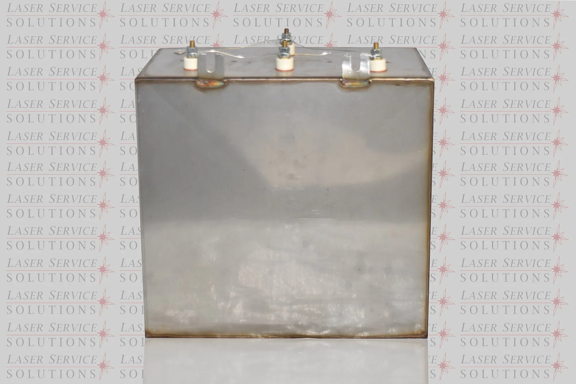 A large, metallic, rectangular Candela MGY Capacitor Bank with four white-capped fittings on top, placed on a reflective surface. The background features repeated Laser Service Solutions logos.