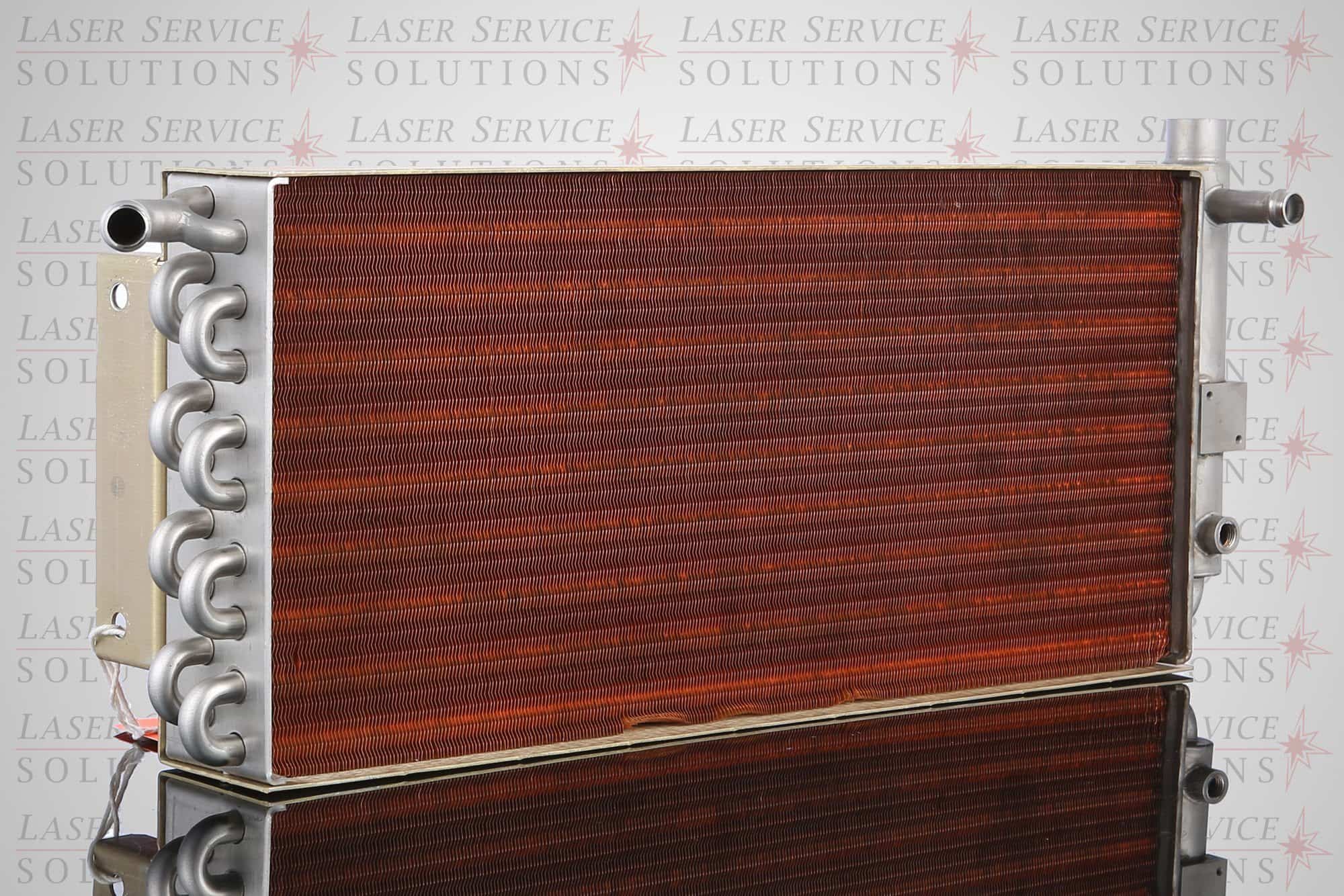 The Elite Heat Exchanger, featuring a rectangular metal design with copper coil fins and multiple pipe connections, stands on a reflective surface with “Laser Service Solutions” faintly visible in the background.