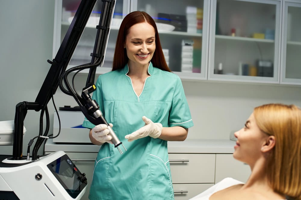 A woman in a plaid shirt looks down thoughtfully while another woman gently rests a hand on her shoulder in a comforting gesture, perhaps offering support as they discuss options like medical laser treatments.